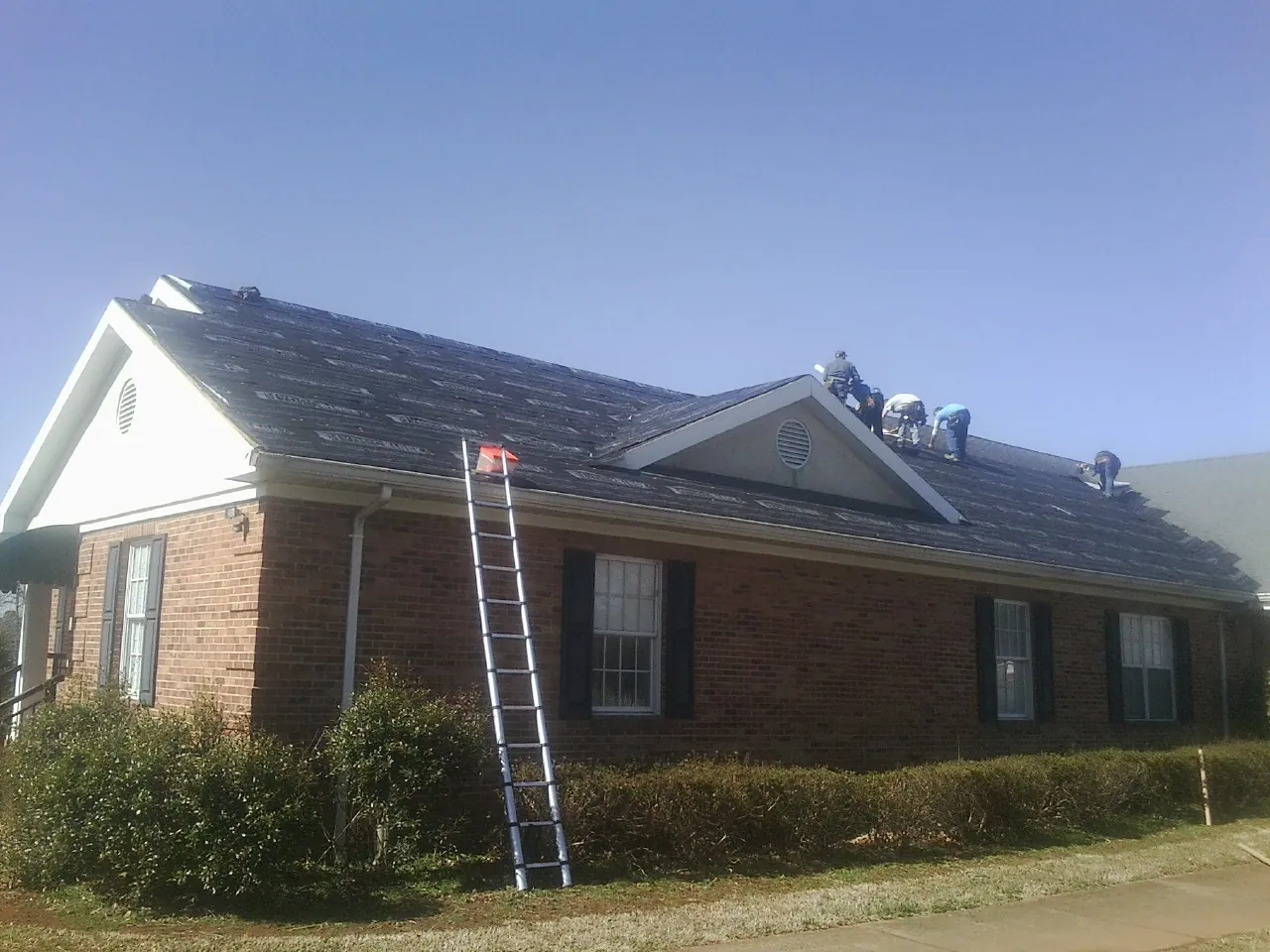 Crew tearing shingles off a brick ranch roof with a ladder set — Metal Roof Repair work by our crew in New Roads