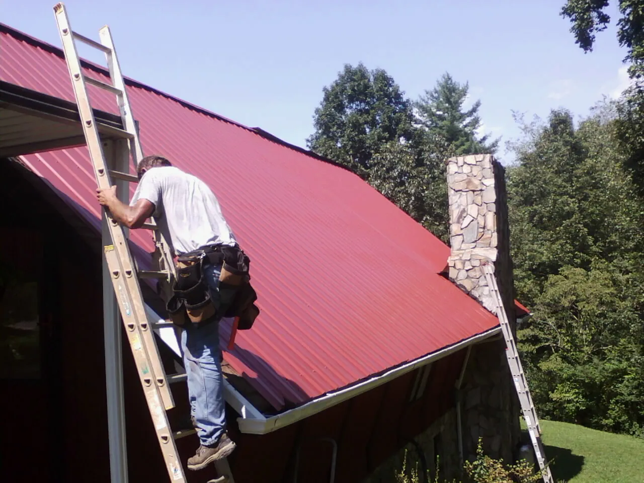 Roofer on a ladder beside a red metal roof and stone chimney — Soffit & Fascia Repair by Roofing Brothers in New Roads